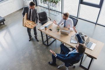 high angle view of young businessman holding pizza in box and looking at coworkers in office