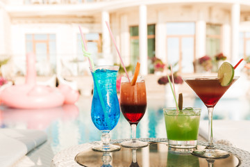 Photo of four cocktail drinks in glasses at table near hotel swimming pool, during sunny summer day