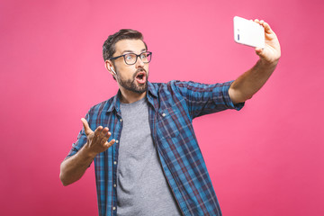 I love selfie! Handsome young man in shirt holding camera and making selfie and smiling while standing against pink background. Listening music with headphones.