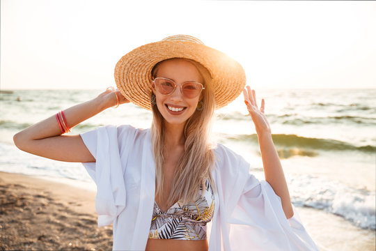 Image of gorgeous blonde woman 20s in summer straw hat and sunglasses smiling, while walking at sea coast