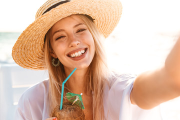 Photo of happy young woman 20s in summer straw hat laughing, and drinking cocktail while taking selfie on beach during sunrise