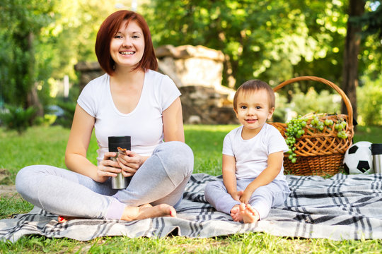 Happy Mother And Little Son Together On Picnic In Park. Mom And Toddler Sit Barefoot, Woman Is Keeping Tumbler Mug And Snack. Basket With Grapes And Ball In Background. Family Healthy Leisure Concept
