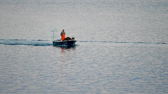 Old traditional fisherman in Croatia on a small wooden boat returning to harbor after catching fish, slow motion shot. Boat is small and in blue color, the fisherman wearing protective orange clothing