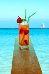 The glass with soft strawberry drink is on a wooden board directly above the sea in clear day. The beautiful view of the sea opens.