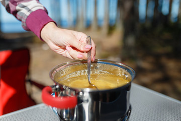 Girl traveler prepares food on portable gas stove, on a folding table on the background of camping in forest. Women's hands interfere with a spoon food in a pot on a gas burner (camping stove).