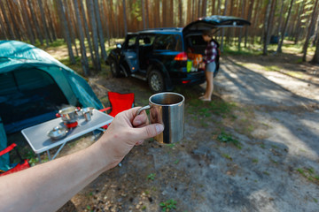 The man's hand holds an iron mug with a warm drink on the background of the table and chairs, campfire, a tent and an SUV in camping. Family vacation, concept of traveling by SUV or car in the wild.