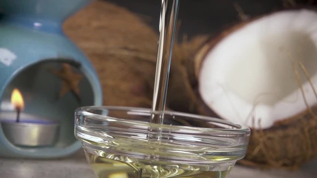 Slow motion natural coconut oil pour a thin stream into a bowl on the background of coconut halves and aroma lamps with a candle