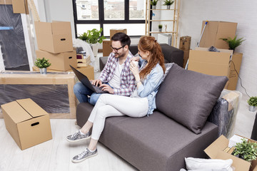 Fototapeta na wymiar Young couple in new home with notebook sitting on sofa and planning with unpacked boxes in background