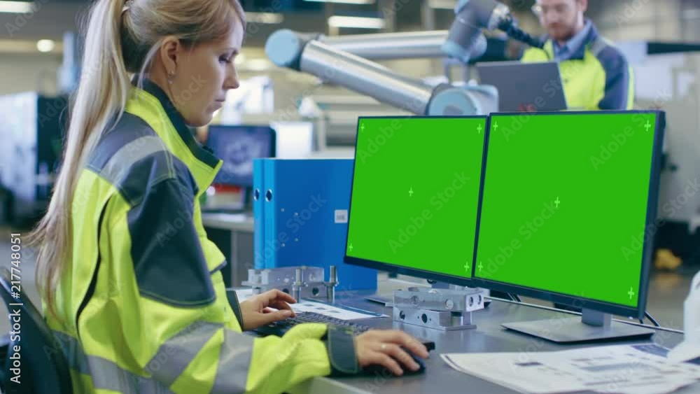 Poster At the Factory: Female Mechanical Engineer Works on Personal Computer with Two Green Mock-up Screen Displays, Male Automation Engineer Works in Background. Shot on RED EPIC-W 8K Helium Cinema Camera.