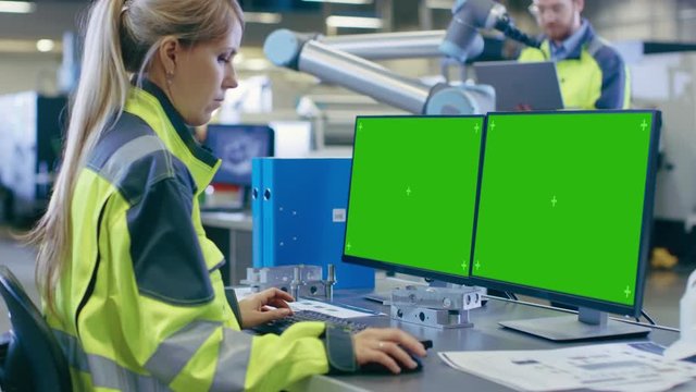 At the Factory: Female Mechanical Engineer Works on Personal Computer with Two Green Mock-up Screen Displays, Male Automation Engineer Works in Background. Shot on RED EPIC-W 8K Helium Cinema Camera.