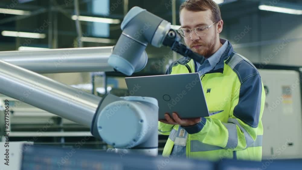 Poster at the factory: automation engineer uses laptop for programming robotic arm. new era in automatic ma