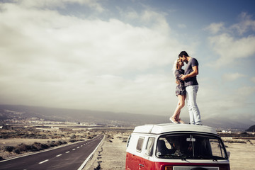 couple of caucasian man and woman people in love standing over the roof of a classic van vintage kissing and hugging. long way road on the side for travel and destination concept
