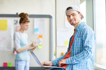 Fototapeta premium Happy boy in casualwear looking at you while typing and working in the net in classroom of college