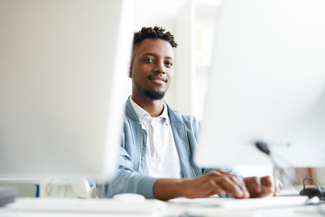 African-american young it-manager looking at you while sitting between two monitors and working...
