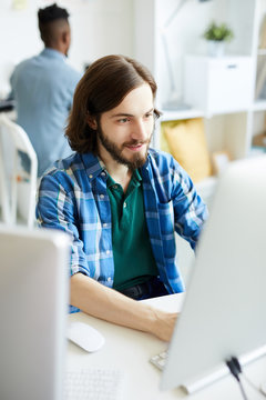 Young bearded man in casualwear sitting in front of computer monitor and carrying out developing work