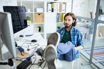 Young relaxed it-manager sitting on chair with his legs on desk and looking at computer screen in...