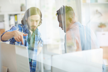 One of young it specialists pointing at computer monitor while explaining his colleague data on screen