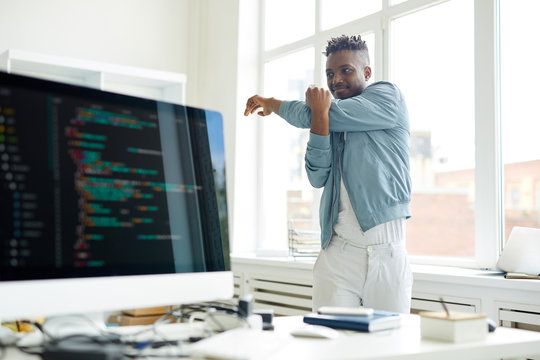 Young it-manager in casualwear standing between window and his desk with computer and doing exercise for arms