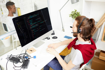 Young it-manager in headset sitting in front of computer monitor, talking to clients and working...