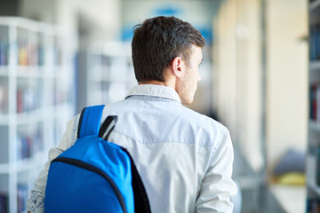 Rear view of pensive boy with stubble carrying satchel on back and moving along corridor while...