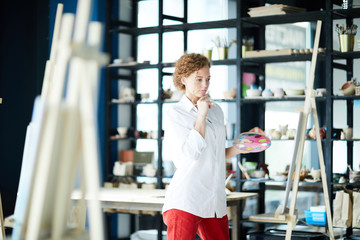 Pensive young artist in casualwear standing in front of her easel and looking at painting in studio of arts