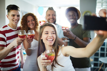 Happy girl with cocktail making selfie of friendly company while having fun at party