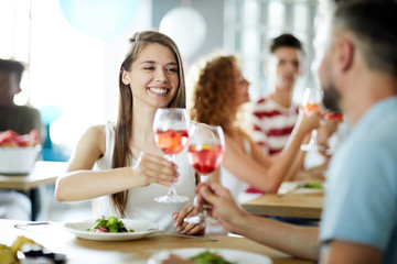 Happy girl and her companion clinking their glasses with drinks while enjoying party by served table