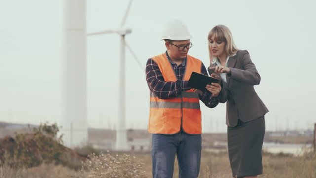 Construction business. Business lady and young man in helmet and reflective vest on windmills background using digital tablet are planning construction. Windmills and the object in the background.