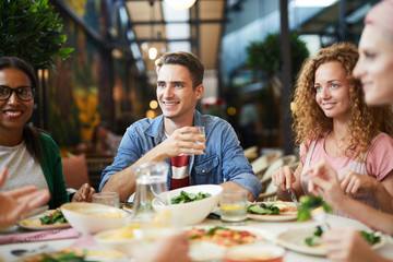 Three young people sitting by served table, having dinner and communicating with friends