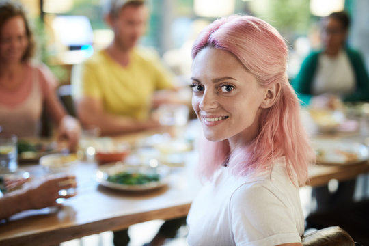Happy young female with pink hair sitting by table and enjoying dinner with friends