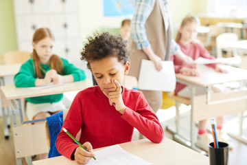 Thoughtful elementary student boy with curly hair sitting at desk and passing test in classroom, he...