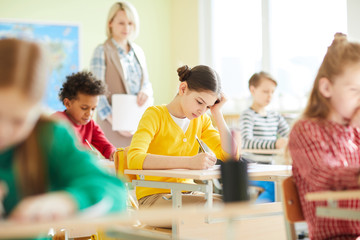 Confused schoolgirl with hair buns sitting at desk and passing test in classroom, she having problem with quiz