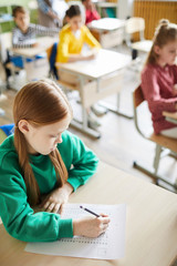 Serious concentrated cute girl in green sweater filling quiz blank while solving task at lesson in...