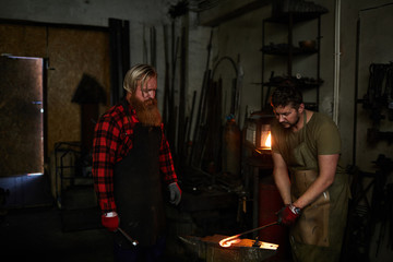 Skilled long-bearded blacksmith in apron teaching young worker to forge metal and controlling his...