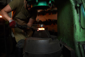 Serious concentrated male manual worker in apron using fire iron while fixing heated detail and...