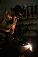 Serious concentrated bearded man in dark goggles using cutting torch while cutting steel detail on anvil in dark workshop, sparks blowing out