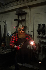 Fototapeta na wymiar Serious handsome blacksmith with long beard wearing apron and checkered shirt holding steel part and cutting it with torch in workshop