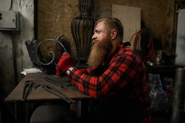 Sad concentrated male blacksmith with long beard checking twisted metal bar made by hands in...