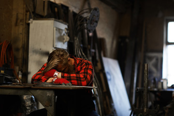 Tired male blacksmith in checkered shirt and gloves sleeping at table and keeping head on arms in...