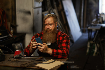 Frowning puzzled bearded blacksmith in checkered shirt using smartphone while checking message and sitting at table with hand tools in workshop