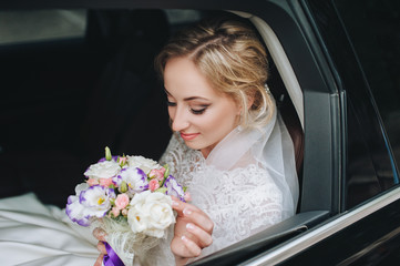 A beautiful bride sits in a black car with a bouquet in her hands. Wedding portrait of a cute bride in the window of a modern car.