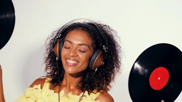 Young beautiful african american woman with headphones in yellow dress enjoying and dancing with vinyl records at white background. Modern trendy girl with afro hairstyle. 4k