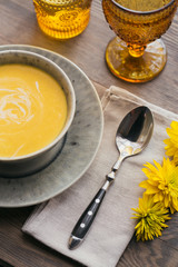Rustic table setting with pumpkin soup, linen napkin, cutlery, ceramic plates, yellow glasses and yellow flowers