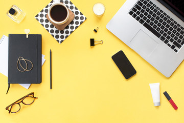 Home office desk. Female workspace with laptop, phone, pencil, candle, women's cosmetic accessories, coffee mug, black diary on yellow background. Flat lay, top view. Fashion blog look. Add your text.