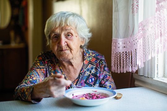 An elderly woman eating soup sitting at a table in the house.
