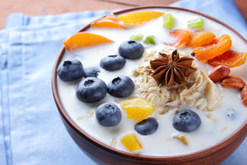Oatmeal porridge with milk, fruits and peanuts on wooden background, healthy porridge with blueberries, banana and peach slices, breakfast on blue napkin
