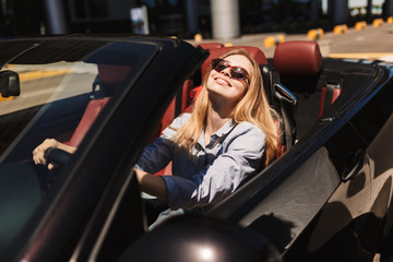 Pretty smiling girl in sunglasses happily looking in camera while driving cabriolet car on city...