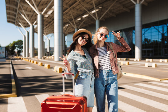 Two beautiful smiling girls in sunglasses happily looking in camera with red suitcase near airport