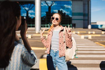 Beautiful girl in sunglasses with backpack on shoulder showing two fingers gesture and sending air kiss while girl near taking photo of her near airport