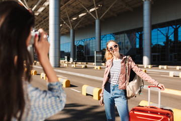Pretty smiling girl in sunglasses with backpack on shoulder and red suitcase happily looking in camera while girl near taking photo of her near airport